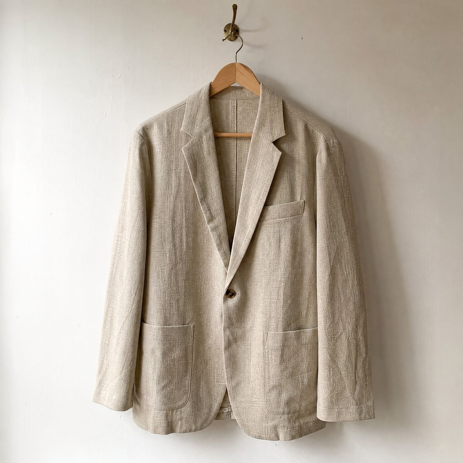 A linen blazer photographed alone on a wooden hanger against a white wall, the starting garment photo.