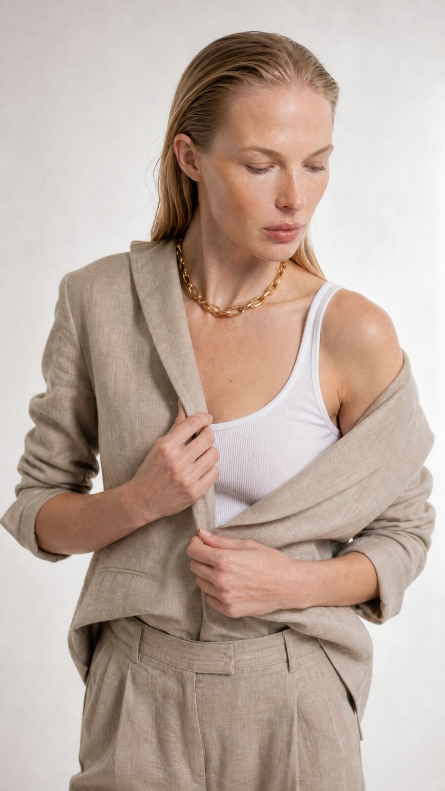 A model in wide linen trousers and a white tank slips a matching linen blazer off one shoulder, against a pale backdrop.