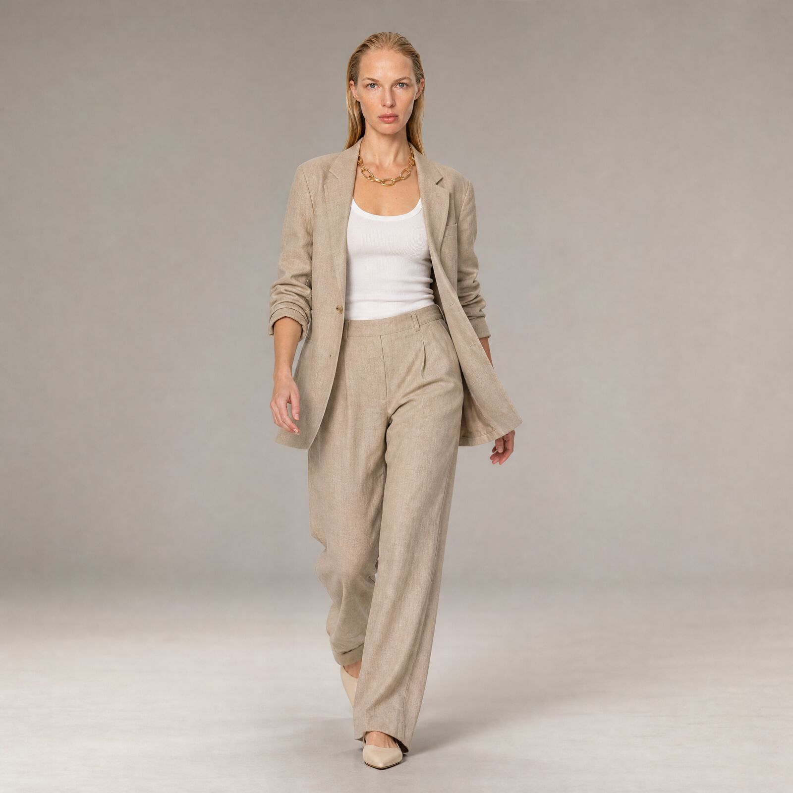 Full-length shot of a model walking toward camera in a linen blazer, white tank, wide linen trousers and nude pointed flats, on a grey studio backdrop.