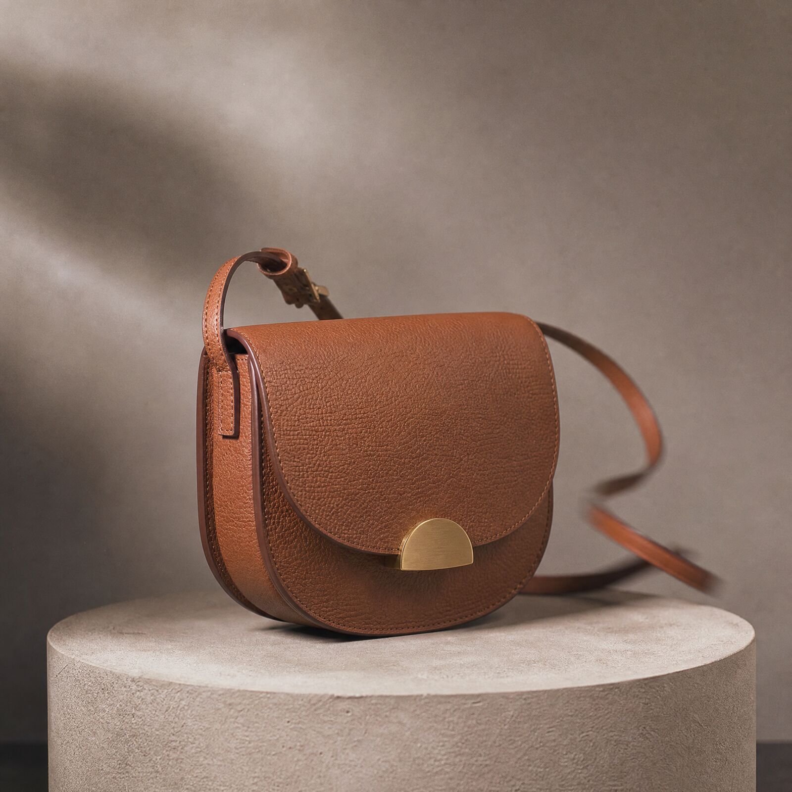 A tan leather saddle bag with a brass half-moon clasp on a round stone plinth, strap looping behind, soft shadows on a warm grey wall.